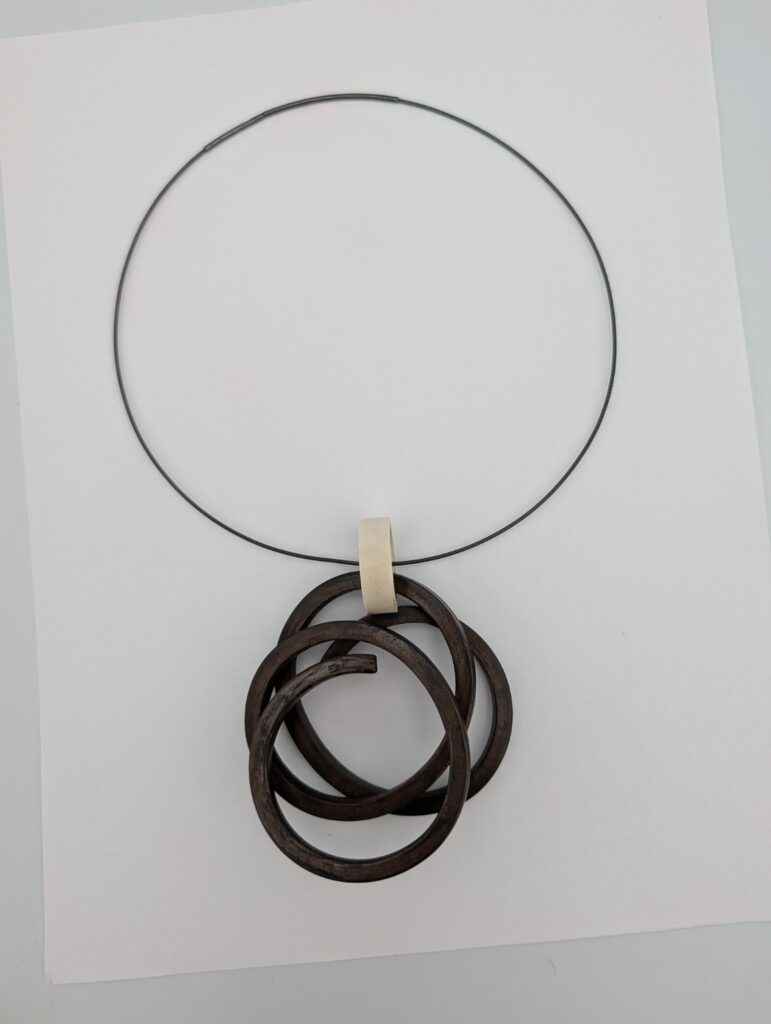 Held Knot Neckpiece by Gustav Reyes, a dark looping bent-wood pendant held by a pale connector on a dark cord.