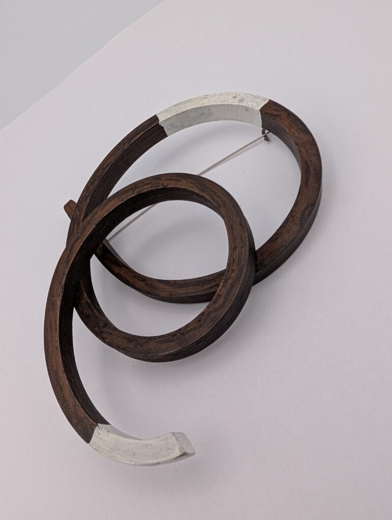 Open Coil Brooch by Gustav Reyes, a curved wooden brooch with an open coil form and metal pin structure.